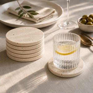 May include: A stack of six beige stone coasters next to a glass of water with a lemon slice. A single coaster is under the glass. A plate with a folded napkin and olives in a small bowl are also in the image.