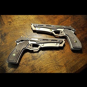 Pode incluir: Duas pistolas de brinquedo de madeira com acabamento em madeira escura, detalhes em latão e o nome "Gavin" gravado em uma. As pistolas estão sobre uma superfície de madeira.
