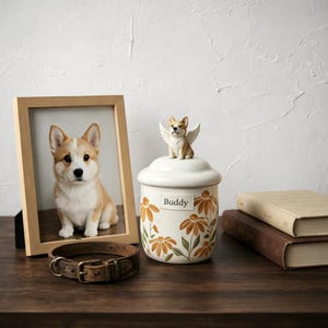 Pode incluir: Uma foto emoldurada de um cão corgi ao lado de uma urna de cerâmica para animais de estimação com uma estatueta de anjo corgi no topo. A urna é decorada com desenhos florais laranja e o nome "Buddy". Uma coleira de cachorro de couro e dois livros também estão presentes.