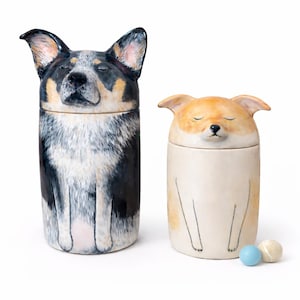 Puede incluir: Dos recipientes de cerámica con forma de perro. Uno es negro, blanco y canela, el otro es crema y canela. Ambos tienen los ojos cerrados y están sentados. Dos pequeñas bolas están frente al perro color crema.