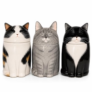 Pode incluir: Três recipientes de cerâmica em forma de gato. O primeiro é branco com marcas pretas e castanhas, o segundo é cinzento e o terceiro é preto com o peito branco. Cada um tem uma tampa em forma de cabeça de gato, com os olhos fechados e uma expressão sorridente.