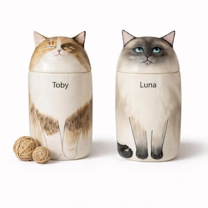 Puede incluir: Dos botes de cerámica con forma de gato, uno con el nombre "Toby" y otro con "Luna". El bote de Toby es de color crema con marcas marrones y tostadas, y el bote de Luna es de color crema con marcas grises y ojos azules. Dos pequeñas bolas de cuerda están junto al bote de Toby.