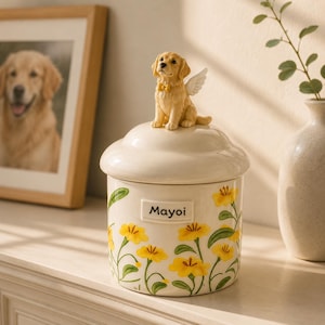 Op de afbeelding: Een crèmekleurige keramische pot met een deksel bekroond door een golden retriever-figuurtje met kleine witte engelvleugels. De pot is versierd met gele bloemen en groene stengels, en een rechthoekig label toont de tekst Mayoi.