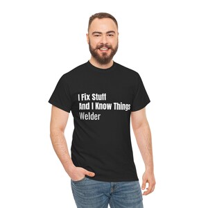 May include: Black crew neck t-shirt featuring the white text: "I Fix Stuff And I Know Things Welder". The person wearing the shirt is also wearing blue jeans.