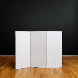 May include: A white, three-panel folding screen stands on a hardwood floor against a black wall. The screen is made of three rectangular panels connected by hinges, allowing it to stand upright. The room has a minimalist aesthetic.