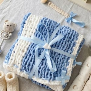 Könnte beinhalten: Ein Baby-Geschenkset mit einer blau-weißen Strickdecke, die mit einem blauen Band gebunden ist. Das Set enthält einen Schnuller, eine Rassel, Babyschuhe und ein Buch mit dem Titel "The Plover's Children's Story".