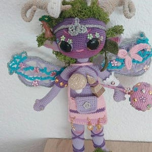 Könnte beinhalten: Eine gehäkelte violette Puppe mit großen schwarzen Augen, geschwungenen Hörnern und grünen Moosakzenten. Sie hat durchscheinende Flügel mit blauen und rosa Blumendetails, einen rosa Rock mit Anhängern und einen passenden Stab mit Perlen.