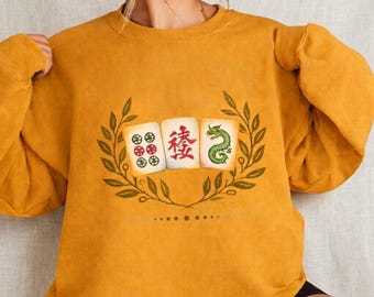 May include: Mustard yellow sweatshirt featuring a Mahjong tile graphic. The graphic includes three tiles with symbols and Chinese characters, surrounded by a green laurel wreath. The sweatshirt is a solid color and has a crew neck.