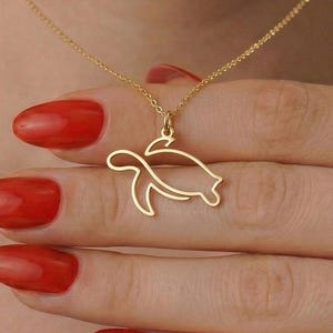 Peut inclure: Un collier doré avec un pendentif en forme de tortue. Le pendentif est un contour minimaliste d'une tortue. Le collier est tenu par une personne avec du vernis à ongles rouge.