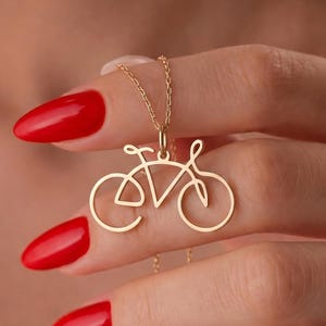 May include: A gold bicycle pendant necklace. The pendant is a minimalist outline of a bicycle. The necklace is held by a person with red nail polish. The chain is delicate and thin.