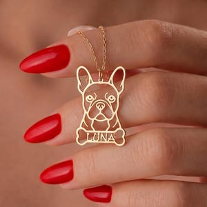 Może przedstawiać: Złoty naszyjnik z wisiorkiem przedstawiającym buldoga francuskiego. Wisiorek to wycięta sylwetka twarzy i ciała psa, z imieniem "LUNA" wpisanym na elemencie w kształcie kości. Naszyjnik trzymany jest przez osobę z czerwonym lakierem do paznokci.