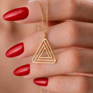 Può includere: Una collana color oro con un ciondolo a forma di triangolo impossibile. Il ciondolo è sospeso a una sottile e delicata catenina color oro. Il design geometrico è composto da linee interconnesse che formano una figura triangolare con il centro aperto.