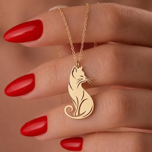 Puede incluir: Un collar de tono dorado con un colgante en forma de gato sentado estilizado de perfil. El colgante cuelga de una delicada cadena dorada. El diseño del gato incluye detalles recortados en las orejas, los bigotes y la curva de la cola.