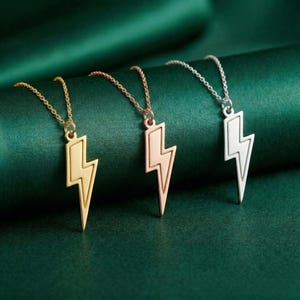 May include: Three necklaces with lightning bolt pendants. One is gold, one is rose gold, and one is silver. The pendants are on delicate chains. The background is a dark green fabric.