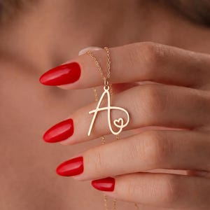 May include: A gold-coloured necklace featuring the letter "A" in a cursive font, with a small heart detail. The pendant hangs from a delicate chain. The image shows the necklace against a blurred background.