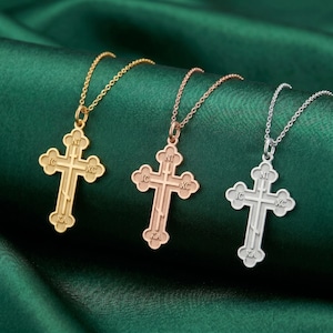 Könnte beinhalten: Drei Anhänger-Halsketten mit orthodoxen Kreuzen in Gold, Roségold und Silber. Jedes Kreuz hat ein detailliertes Design mit religiösen Inschriften und hängt an einer zarten Kette. Die Halsketten werden auf einem dunkelgrünen Stoff präsentiert.