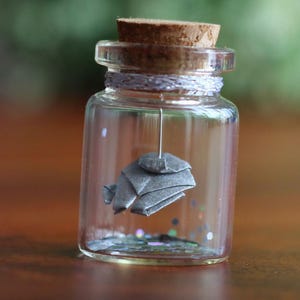 Puede incluir: Una pequeña botella de vidrio transparente con tapón de corcho que contiene un elefante de origami metálico plateado suspendido de un hilo fino. La base de la botella está llena de pequeñas partículas de purpurina iridiscente que reflejan la luz.