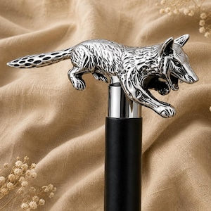 Puede incluir: Un bastón con un mango de metal plateado en forma de lobo saltando con textura de pelaje detallada y la boca abierta. El mango está montado sobre un eje negro cilíndrico y liso con un collar de metal pulido de color plateado.