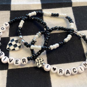 May include: Three beaded bracelets with a racing theme. The bracelets are black and white, with the words "YOUR DRIVER" and "I ❤️ RACING" spelled out in white letter beads. One bracelet has a checkered heart charm.