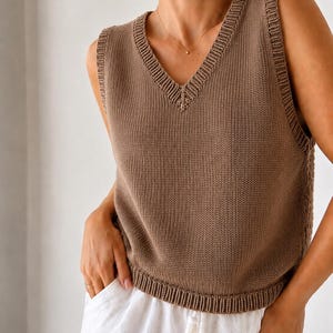 May include: A brown sleeveless knit sweater vest featuring a V-neckline with ribbed trim along the collar, armholes, and hem. The garment is constructed with a standard stockinette stitch pattern and is worn over white trousers.