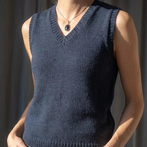May include: A navy blue knitted vest with a V-neck. The sleeveless top is made of a ribbed knit fabric. A dark pendant hangs from a gold chain around the neck. The vest is a solid color.