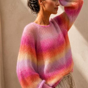 Könnte beinhalten: Ein langärmeliger, flauschiger Strickpullover mit einem horizontalen Farbverlauf aus leuchtenden rosa, orangefarbenen und hellvioletten Streifen. Das Kleidungsstück hat einen Rundhalsausschnitt, Raglanärmel und gerippte Bündchen, kombiniert mit einer beigen Hose.