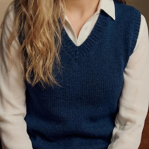 May include: A navy blue knitted V-neck vest. The vest is worn over a white collared shirt. The image shows the upper body of a person with long blonde hair. The vest is a solid color and appears to be made of a soft material.