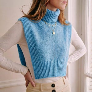 Puede incluir: Un chaleco de cuello alto de punto azul claro. El chaleco sin mangas tiene un dobladillo acanalado. La modelo lleva una camisa blanca de manga larga y un collar dorado. El chaleco es una pieza elegante para combinar.