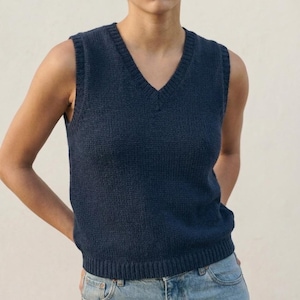 May include: A navy blue, sleeveless sweater vest with a V-neck. The knitted vest has a ribbed hem and a classic, casual style. The model is wearing the vest with blue jeans.