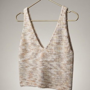 May include: A sleeveless knit tank top featuring a deep V-neckline and a ribbed texture. The garment is crafted from a variegated yarn blend in soft shades of cream, beige, and light brown, creating a subtle mottled appearance throughout the fabric.