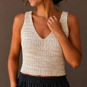 May include: A sleeveless, cropped knit tank top in a heathered beige and cream color palette. The garment features a deep V-neckline and a vertical ribbed texture throughout the bodice, ending in a straight hemline that sits at the waist.