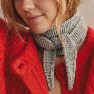 May include: A close-up of a gray knitted scarf tied around the neck. The scarf is made of a thick, textured yarn. The person is wearing a bright red cardigan with a large button.