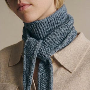Könnte beinhalten: Ein schieferblauer Strickschal mit gerippter Textur um den Hals und einem festen, dichten Strickmuster an den herabhängenden Enden. Der Schal ist vorne einfach geknotet und zeigt die gleichmäßige blaue Garnfarbe sowie die detaillierte Strickarbeit.