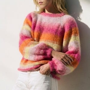 May include: A colorful, striped sweater in shades of pink, orange, and yellow. The sweater has a round neckline and long sleeves with a slightly puffed design. The model is wearing white jeans.