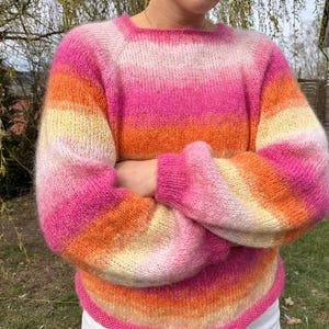 Op de afbeelding: Een handgebreide trui met een levendige kleurovergang van roze, oranje en geel. De trui heeft een ronde hals en lange mouwen. De onderkant en manchetten zijn felroze. De trui is gemaakt van zacht, getextureerd garen.