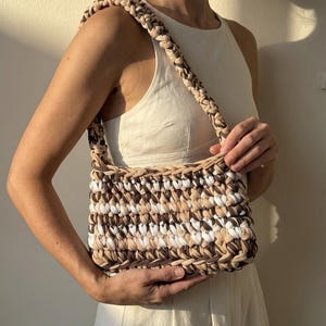 Könnte beinhalten: Eine rechteckige Handtasche mit geflochtenem Riemen, hergestellt aus einer Mischung aus weißem, beigem und braunem Stoff. Die Tasche wird von einer Person gehalten, wodurch die gewebte Textur und die neutrale Farbpalette hervorgehoben werden. Das Design der Tasche ist schlicht und stilvoll.