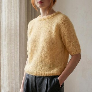 May include: A short-sleeved, pale yellow knit jumper featuring a crew neckline and a fuzzy, textured yarn finish. The garment has ribbed detailing at the collar, sleeve cuffs, and hemline, providing a structured fit for this soft, light-coloured pullover.