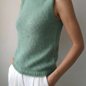 May include: A sage green sleeveless knit top featuring a boat neckline and a ribbed hem. The garment is constructed with a stockinette stitch pattern and is paired with white trousers. The design is simple, focusing on the texture of the knit fabric.