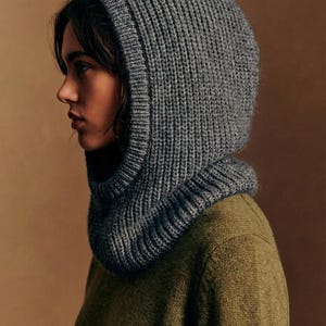May include: A grey knitted hood and cowl. The hood, made from a thick, ribbed knit, covers the head and neck, while the cowl sits around the neck. The person in the image is wearing an olive green jumper.