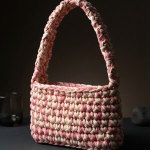 Könnte beinhalten: Eine rechteckige Handtasche aus dickem, geflochtenem Stoffgarn in einem melierten Muster aus zartem Rosa und Creme. Die Tasche verfügt über einen einzelnen integrierten Henkel, einen strukturierten Boden und ein durchgehendes Häkelmuster.