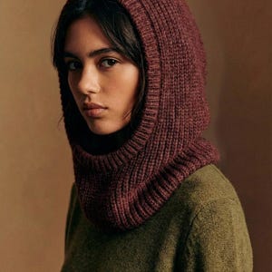 May include: A burgundy knitted hood with a ribbed texture. The hood is worn over a green jumper. The hood covers the head and neck, with an opening for the face. A cosy winter accessory.