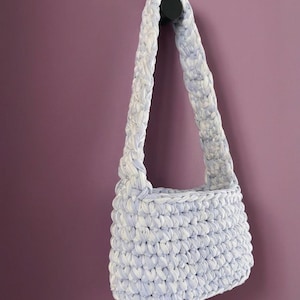 May include: A shoulder bag crafted from thick, braided fabric yarn in a variegated pattern of light blue and white. The bag features a rectangular body with a single integrated strap, showcasing a consistent crochet-style texture throughout.