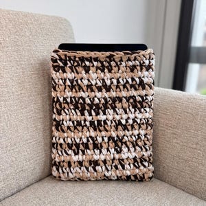 Puede incluir: Una funda rectangular para tableta hecha a mano con ganchillo en una mezcla de marrón, beige y blanco. La funda está hecha de hilo grueso, creando una apariencia texturizada. El borde superior es de color beige liso. La funda está sobre un sillón beige.