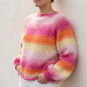 May include: A crew neck sweater in a gradient of pink, orange, and yellow. The sweater has long sleeves with ribbed cuffs and a ribbed hem. The yarn has a soft, fuzzy texture. The model is wearing white pants.