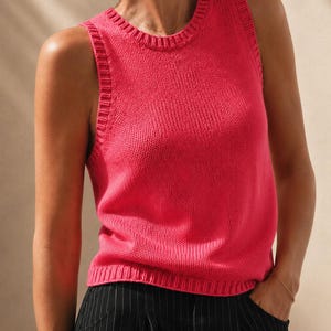 May include: A bright pink sleeveless knit tank top featuring a ribbed crew neckline and ribbed armholes. The garment has a straight hem with a ribbed finish and is paired with black trousers that feature a subtle white pinstripe pattern.