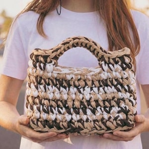 Könnte beinhalten: Handgefertigte Häkeltasche aus braunen, beigen und weißen Garnen. Die Tasche hat einen geflochtenen Griff und eine rechteckige Form. Die Textur des Garns ist sichtbar und erzeugt ein einzigartiges, strukturiertes Aussehen.