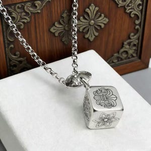 May include: A silver-toned necklace with a cube-shaped pendant. The pendant features engraved floral and ornate designs on each face. The chain is a linked design. The background is a wooden cabinet.