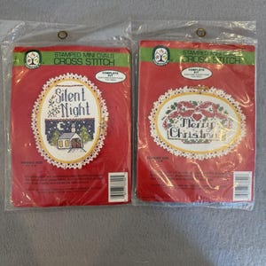 May include: Two Colortex stamped mini oval cross stitch kits in clear plastic packaging. The left kit features a Silent Night design with a church, while the right kit displays a Merry Christmas design with a red bow. Both include a 4 inch by 6 inch wood hoop.
