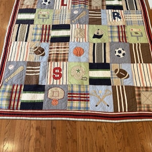 May include: A patchwork quilt with squares featuring sports-themed designs. The quilt includes images of a football, baseball, basketball, and a baseball cap. The border is red and white striped.