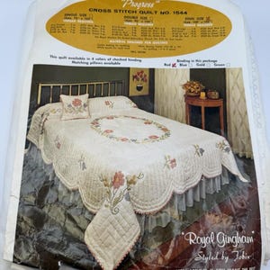 May include: A vintage cross-stitch quilt kit, featuring a floral design on a cream-colored background. The kit includes a quilt, pillow, and a matching bed skirt. The packaging reads "Royal Gingham" and "Progress".